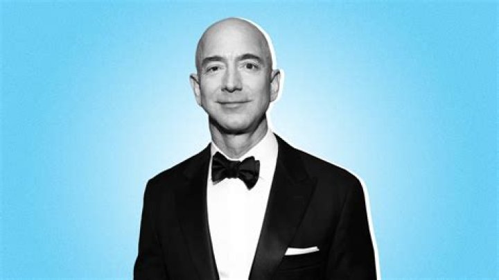 Jeff Bezos in a black and white suite, purple checkered tie, and smiling with his mouth closed. 